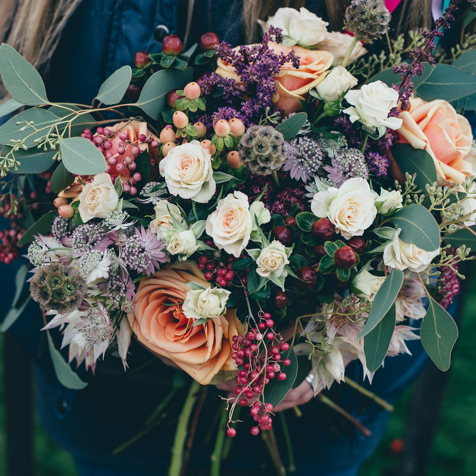 A wild hand-tied bouquet of peach roses, berries and eucalyptus held in both hands